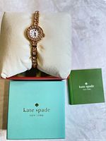 Wristwatch on white cushioned pillow inside open red interior box, green instruction booklet beside next to light turquoise Kate Spade New York outer box