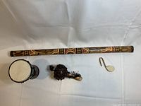 Five folk instruments displayed on white cloth: hand-painted bamboo rainstick, rope-tensioned Jamaican mini drum, small gourd maraca with beads, carved bone musical note ornament, rustic gourd flute