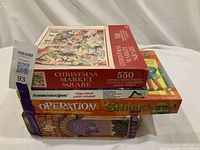Stacked view of the Christmas Market Square puzzle box on top, Operation Shrek Edition game box below, and other games stacked beneath