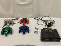 Black Nintendo 64 console, four original Nintendo 64 controllers (grey, red, green, blue), Controller Pak and Jumper Pak accessories, power and AV cables on a white cloth background.