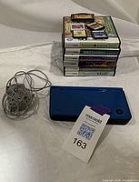 Image showing blue Nintendo DSi XL console closed with wall charger coiled beside it, stacked games and loose cartridges visible in background.