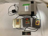 Nintendo Entertainment System console with original rectangular controller, power adapter, AV cable, and three game cartridges (The Legend of Zelda gold cartridge, Dragon Quest, Super Mario Bros. 3) visible. Items laid out on cloth surface.