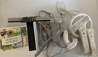 Photo showing white Nintendo Wii console, sensor bar, power adapter, AV cables, 2 Wii Remotes with silicone sleeves, 2 Nunchuks, and Wii steering wheel