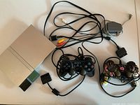 Top-down view of PlayStation 2 console with connected power and AV cables, original Sony controller, and Simpsons-themed third-party controller all laid out on flat surface.