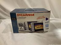 Front view of the sealed product box showing Sylvania 7" Dual Screen Mobile DVD System with extra 7" LCD monitor included.