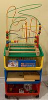 Photo of the colorful three-tier rolling storage cart holding the large wooden wire bead maze on top, visible shelves below contain boxes and toys.