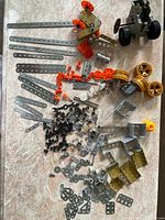 Top-down view of an assortment of vintage Meccano-style metal construction parts including flat strips, angle brackets, orange gears, yellow wheels, partially assembled models, and various hardware pieces arranged on a marble surface.