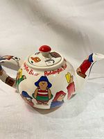 Side view of cream ceramic teapot with colorful Paddington Bear themed illustrations around the body, handle, and spout, including a red lid knob.