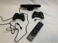 Two black wireless Xbox 360 controllers, one missing its battery pack, Xbox 360 Kinect sensor bar, and Xbox 360 cooling fan attachment with visible dual fans.