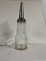 Full view of clear glass oil bottle with attached metal spout and screw cap on top, showing front embossed brand name and location.