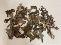 Top-down view showing a scattered assortment of approximately 60 antique and vintage metal keys on a white surface, indicating age with rust and tarnish.