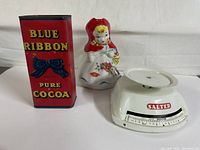 Three item lot showing Blue Ribbon Pure Cocoa tin, Red Riding Hood ceramic pitcher, and Salter kitchen scale together.