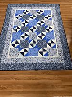 Full view of quilt laid flat on floor showing pattern of patchwork squares in various blue fabrics with white and navy pinwheel accents, and floral border in dark and light blue.