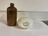 Brown DBC ceramic hot water bottle with black logo alongside white Grimwades 'The Mecca' hot water bottle base showing printed text and registration numbers