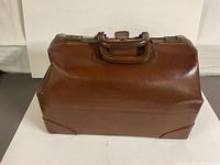 Front and top view of brown leather doctor’s bag with brass clasps and wooden handle, on white surface.