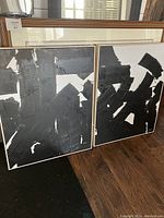 Two paintings side by side leaning against wall with other frames behind