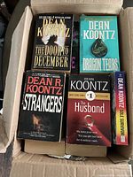 Books in a cardboard box showing titles The Door to December, Dragon Tears, Strangers, The Husband, Sole Survivor by Dean Koontz.