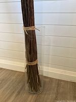 Tall clear glass vase with long bundled brown twigs tied with string inside, standing on a wooden floor against white wall.