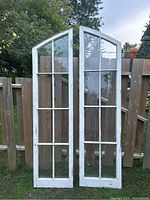Full view of two white wooden arched doors with multiple glass panes resting against a wooden fence outdoors.