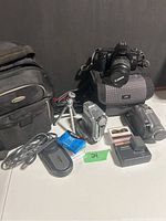 Photo showing Canon EOS 3000 camera on top of JVC camera bag with Panasonic and Samsung video cameras, various cords, tapes, and battery charger in front