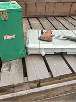 Green metal Coleman storage box with latch, vintage condition, sitting next to Brinkmann camping stove and hatchet with leather blade cover on wooden deck.