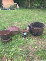 Photo showing all four cast iron items arranged on grass: large cauldron with handle, pot with lid, small pot on stand, and small cauldron.