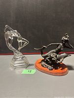 Pair of female statues: the mermaid with dolphin on the right and the clear glass Art Deco female figure on the left against a black background.