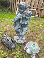 Photo showing the metal children statue with a girl and boy, a steel pig tea light holder with holes for light, and a turtle sprinkler with a number label on its back, all placed on grass.
