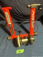 Full view of red metal frame bike trainer with resistance unit and wire cable showing Minoura and Mag 500 branding.