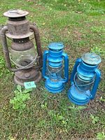 Three lanterns placed on grass; two blue Dietz lanterns and one rusted Beacon lantern.