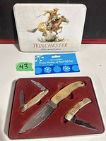 Photo of Winchester 2005 Limited Edition Ersatz Mother of Pearl Gift Set including three knives in a red velvet insert inside a metal presentation tin with cowboy artwork on lid.