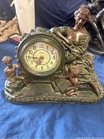 Front view of heavy decorative mantle clock showing clock face, seated woman figurine, two cherub figurines, and ornate base.