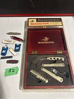 Full view of Winchester deluxe four piece tool set in wooden case and assorted small jack knives and keychain multi-tools arranged around it