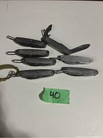 Photo showing seven textured metal pocket tools laid out on white surface, one partially opened displaying blades and tools, with a green label marked '40'.