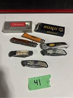 Top down photo showing 8 assorted pocket tools including Victorinox, Ruko, Stubby folding knives laid out on white surface with green tape marked '41'. Two knives are in original boxes.