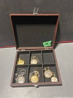 Five pocket watches arranged in a wooden case with six velvet-lined compartments, one compartment empty.