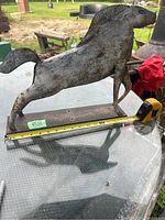 Side view of primitive horse metal sculpture showing its profile and rusted finish on a glass table with measuring tape.