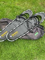Pair of black Yanes Mountain Pass adjustable snowshoes resting on a black carrying bag on grass.