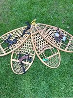 Four wooden snowshoes with woven rawhide lacing and foot straps lying on grass.