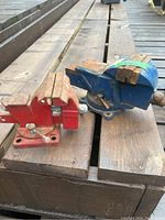 Two metal bench vices placed on a wooden surface, showing their overall condition and mounting bases.