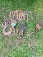 Photo showing three vintage cast iron horse shoes, a large hook with eyelet, two smaller curved hooks and one block and tackle double pulley with hook on grass.