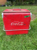 Full front view of the red Coca Cola cooler with closed lid and carry handle on top and side handle for lifting.
