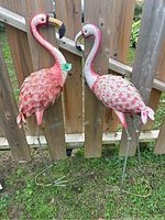 Pair of standing metal flamingo statues against a wood fence on grass, showing their full body front view.