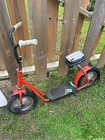 Side view of vintage red two-wheeled scooter with black rubber tires, white handle grips, and checkered seat. Shows overall condition and structure.