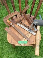 Photo of vintage tools including wooden mallet, metal vice clamp, rusted measuring squares, wooden level, metal ruler, and angle measuring tool displayed on wooden chair
