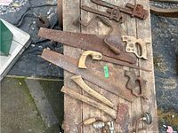 Top-down view of 3 rusty hand saws with wooden handles, 2 pipe wrenches in the upper corner, a wooden hatchet handle and other metal hand tools on a wooden tabletop surface.