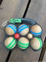 Top view of six wooden pétanque balls, the small red jack ball, all in a black plastic carrying case with a handle. The balls have alternating blue and green stripes.