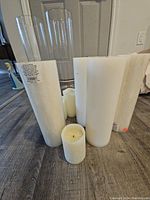 White assorted candles on floor including tall new pillar candles in plastic wrapping and shorter, used round pillar candles