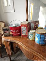 A display of assorted vintage tin cans and a red metal beer bucket on a wooden dresser with mirror, showing the variety of designs and sizes.