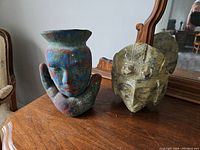 View of two Aztec-style sculptures on a wooden surface in front of a mirror, showing a colorful face held by a hand and a carved stone face mask.